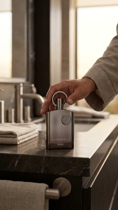 A hand holding the cap of a matte dark gray rectangular perfume bottle on a dark marble bathroom counter.