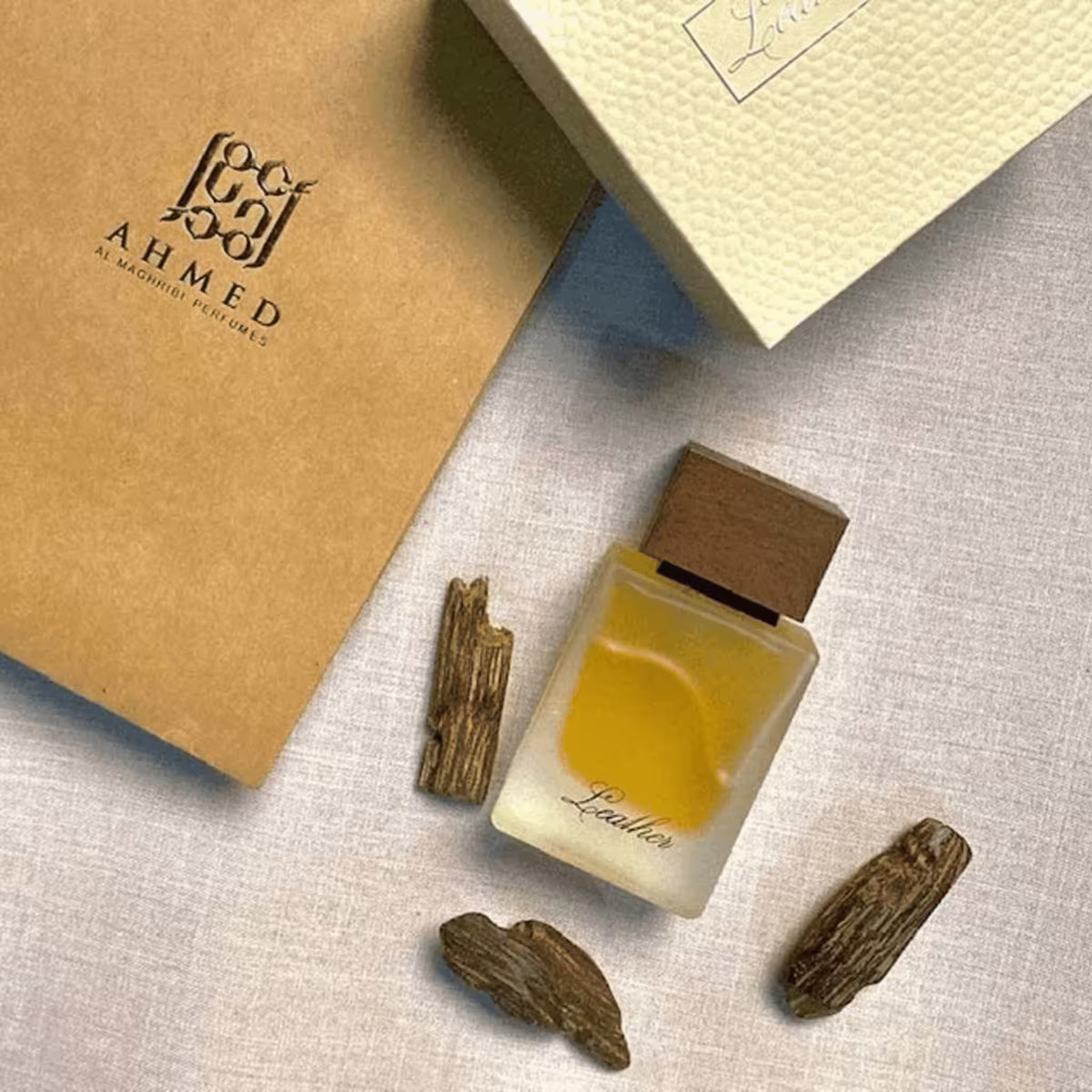 A perfume bottle labeled 'Leather' by Ahmed Al Mahmoud of Perfumes, filled with amber-yellow liquid and topped with a dark wood cap, arranged on a light fabric surface with three wood pieces and branded packaging.