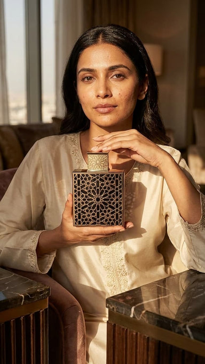 A woman in a cream-colored embroidered kurta holds a square wooden box with intricate geometric cutout patterns, set in a warm, sunlit indoor environment.